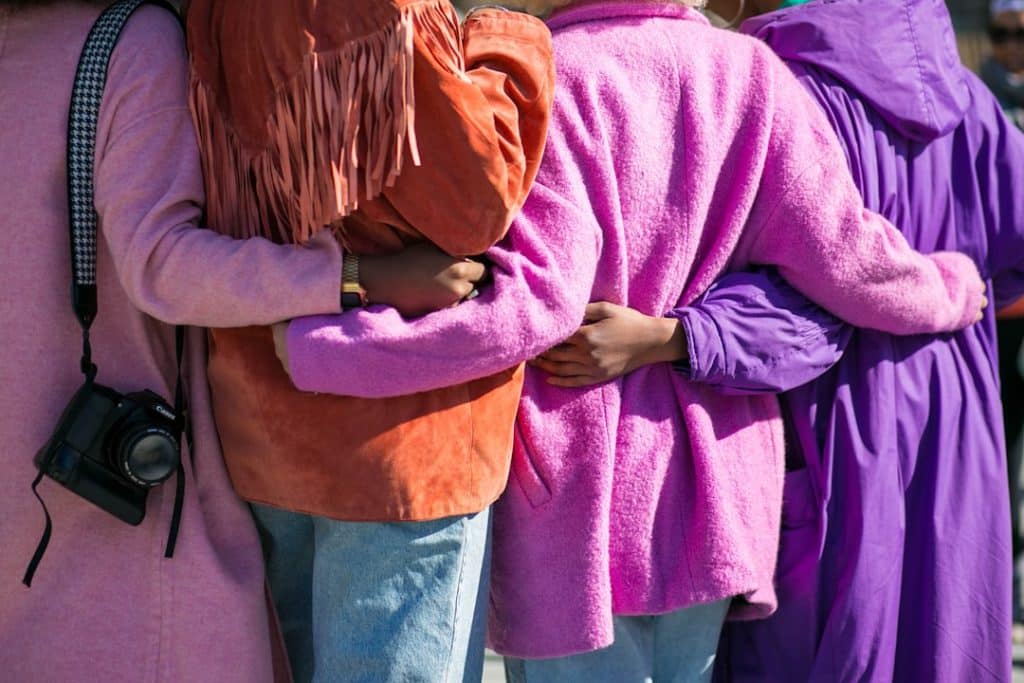 Four individuals stand close together, arms wrapped around each other. They're wearing colorful jackets in pink, orange, and purple hues, with one person holding a camera by their side. The group faces away, evoking a sense of friendship and support.