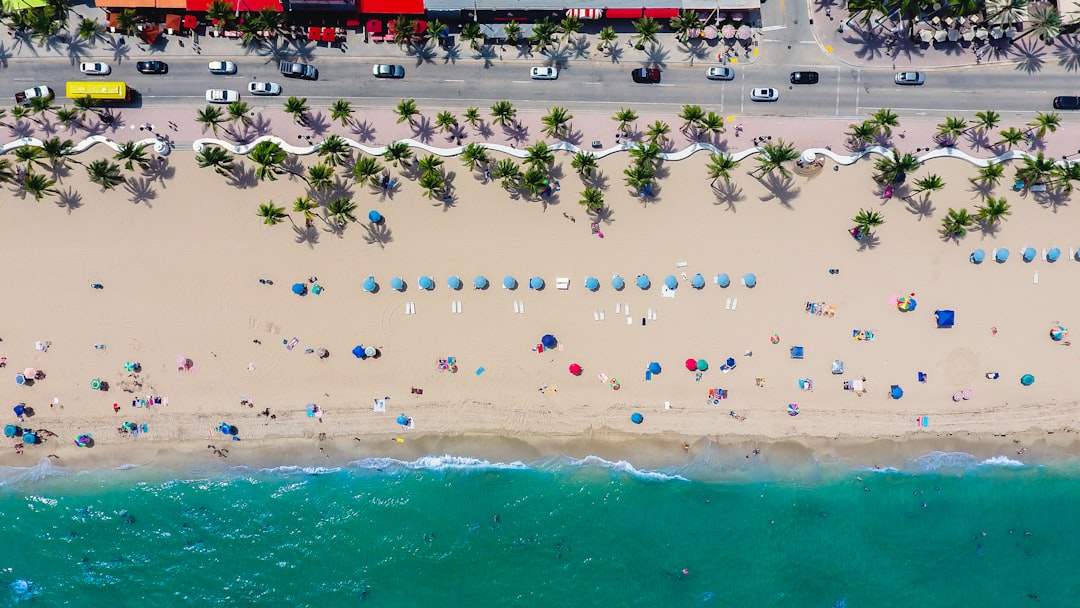 Aerial view of a sandy beach with umbrellas, sunbathers, and a turquoise ocean. A road lined with palm trees runs parallel to the beach with parked cars and buildings. Brightly colored beach towels and umbrellas dot the sand.