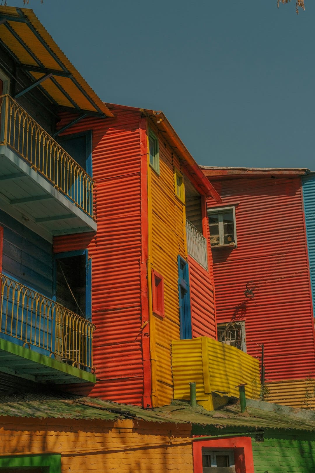 Colorful buildings with corrugated metal siding in shades of orange, yellow, red, and blue, featuring small windows and balconies. The vibrant structures are set against a clear blue sky, creating a lively and cheerful atmosphere.