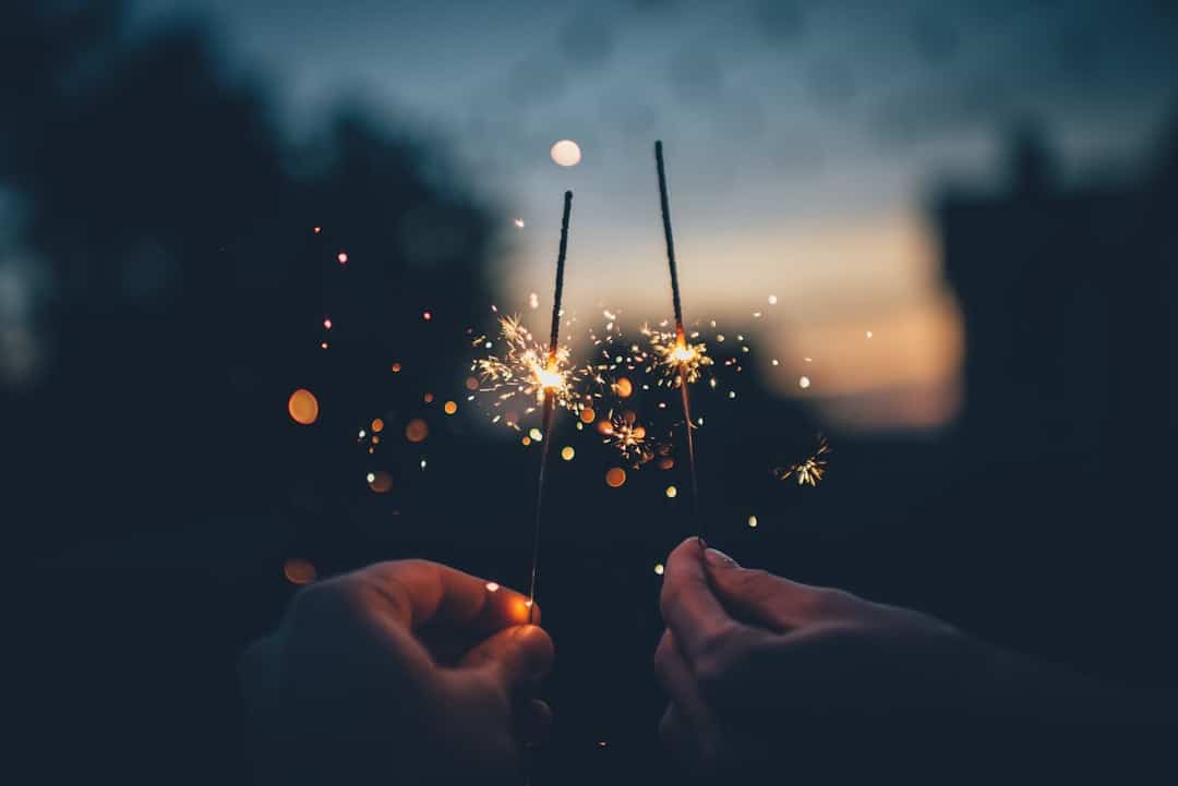 Two hands holding sparklers illuminate the evening sky. The sparklers create a bright, sparkling effect against a dark, blurred background, producing a festive and lively atmosphere.