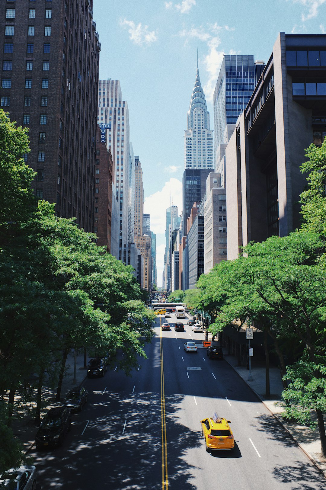 A city street view of New York with tall buildings flanking both sides. A few yellow taxis and cars drive down the avenue. Trees line the sidewalks, and the iconic Chrysler Building is visible in the background under a clear blue sky.