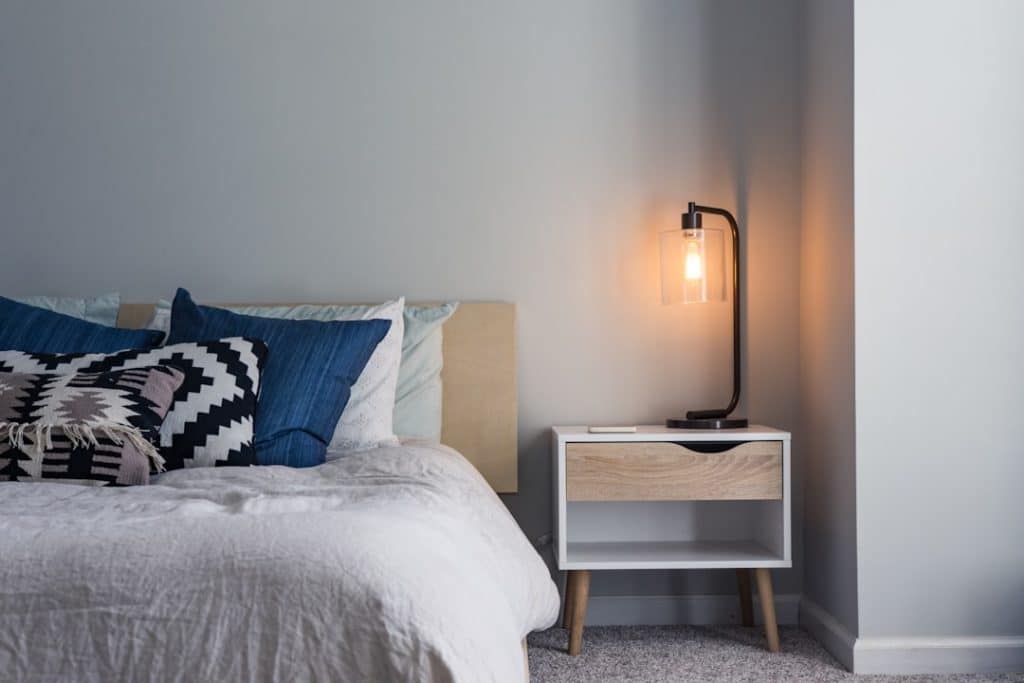 A cozy bedroom features a neatly made bed with blue and black patterned pillows. A modern bedside table with a drawer holds a lit lamp, casting a warm glow. The walls are painted light gray, and a soft carpet covers the floor.