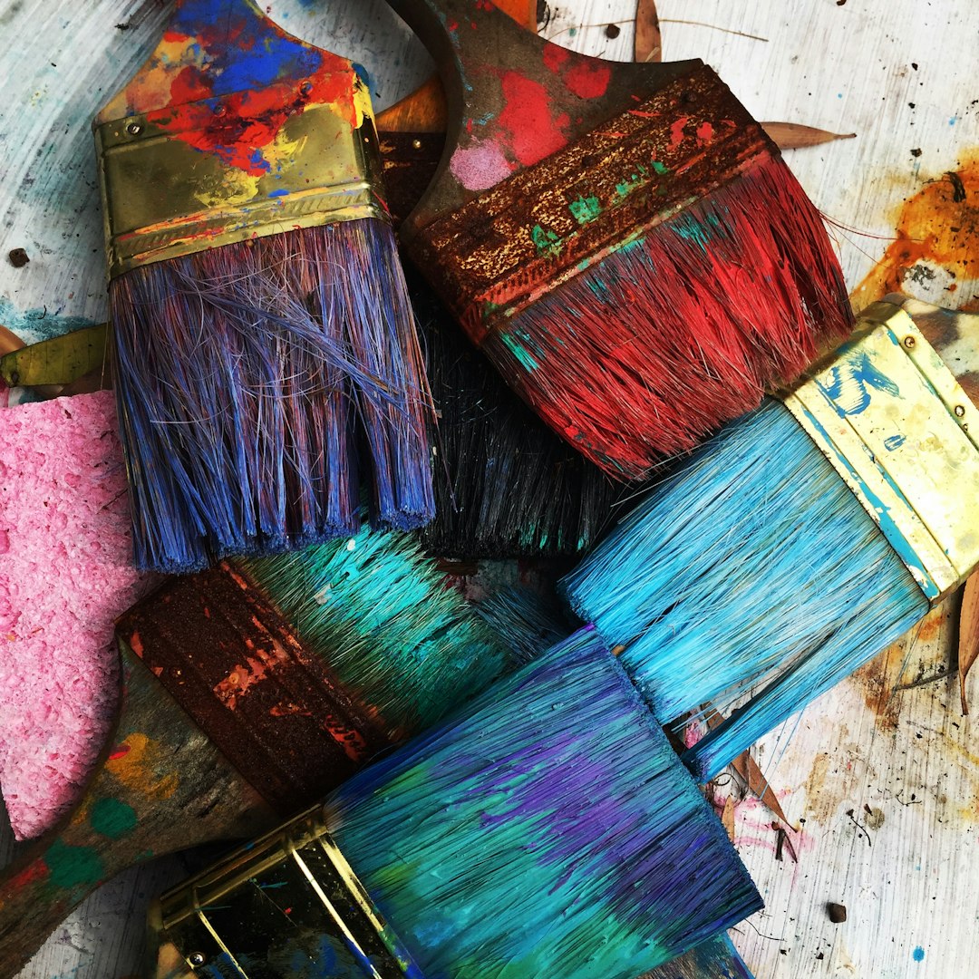 A collection of six paintbrushes with colorful, paint-splattered bristles in shades of red, blue, purple, and teal. The brushes are laid out on a textured surface with visible paint spots.