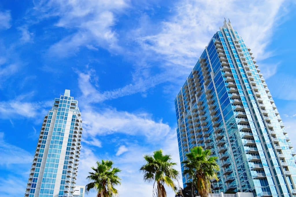 Two tall modern buildings with blue glass exteriors under a bright blue sky filled with wispy clouds. Three palm trees stand in the foreground, adding a touch of greenery to the urban scene.