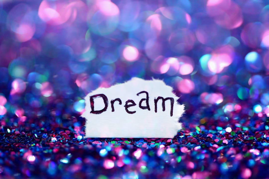 The word "Dream" is handwritten on a torn piece of paper set against a sparkling background of purple, pink, and blue lights, resembling bokeh or glitter.