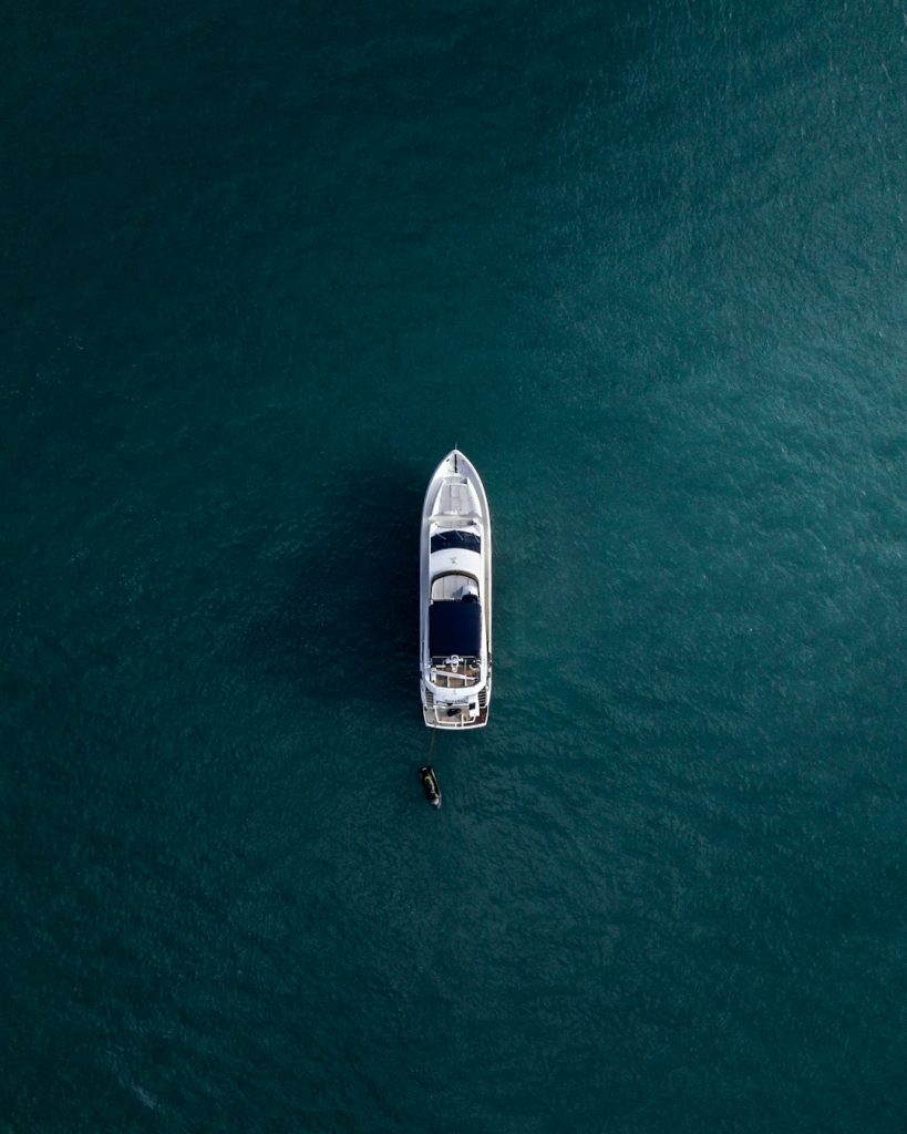 Aerial view of a white yacht with a dark roof floating on clear, deep blue water. A small dinghy is positioned next to the yacht on the left side. The ocean appears calm and expansive, with no visible land around.