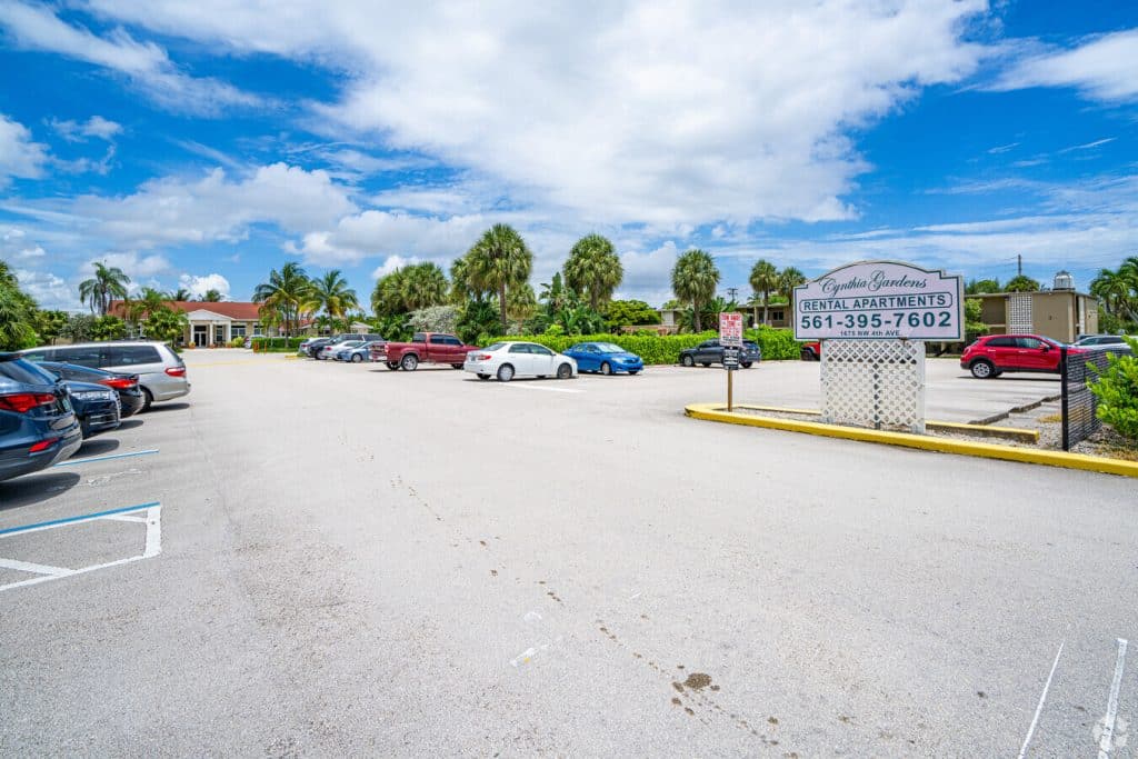 A parking lot with several cars sits under a partly cloudy sky. To the right, a sign reads "Carlton Gardens Rental Apartments 561-395-7602." Palm trees and upscale apartment buildings showcase the charm of Boca Raton in the background.