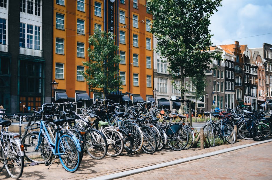 A street scene with numerous bicycles parked along a sidewalk in front of colorful buildings. The background shows a mix of modern and traditional architecture under a partly cloudy sky.