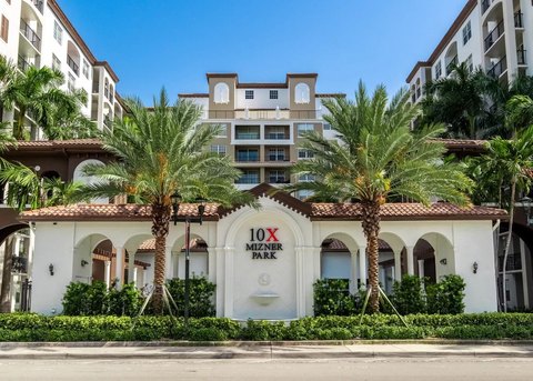 The image shows a residential building named "10X Mizner Park," featuring a white facade with red-tiled roofs and archways. Two large palm trees and green bushes adorn the entrance, under a clear blue sky.