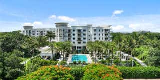 A white, multi-story luxury building is surrounded by lush greenery. In front, there's a large swimming pool flanked by palm trees. The sky is clear and blue, adding to the serene and tropical ambiance.