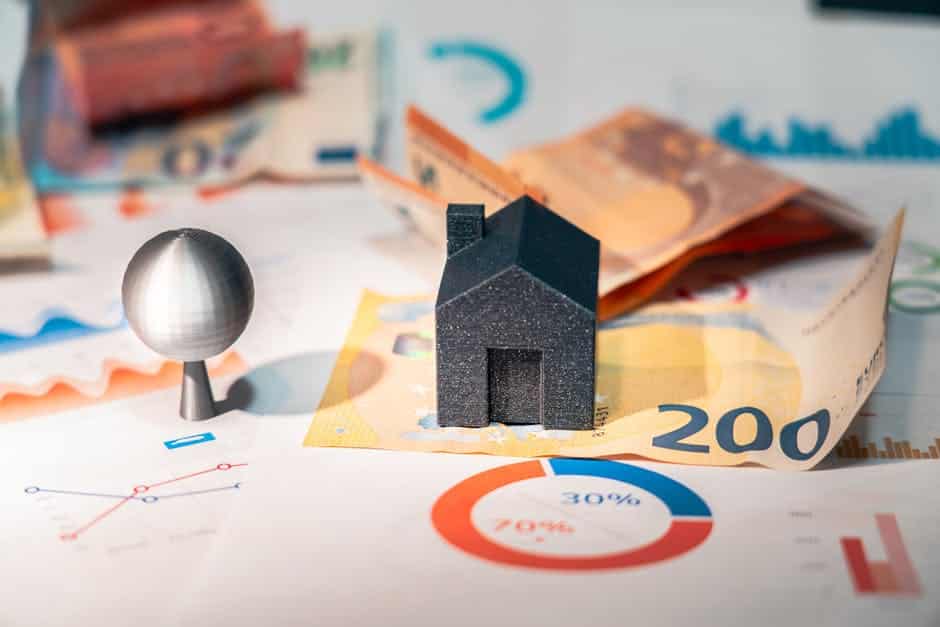 A miniature black house and silver sphere sit on sheets of paper displaying charts and Euro banknotes. The setup represents financial concepts and real estate investments, with colorful graphs and currency suggesting economic analysis.