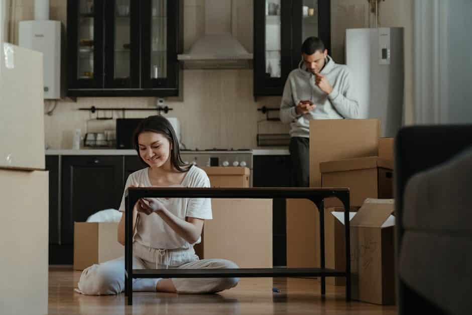 A woman sits cross-legged on the floor assembling furniture, surrounded by cardboard boxes, in a modern kitchen. A man stands in the background checking his phone. Both appear focused on their tasks amidst a moving or unpacking scene.