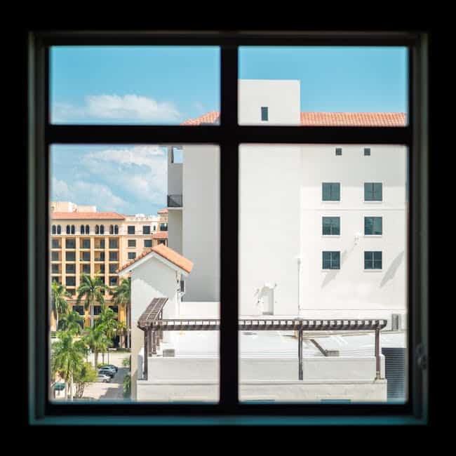 View through a window showing a cityscape with several beige and white buildings, palm trees, and a clear blue sky. The scene is framed by the window's dark grid, dividing the view into four sections.