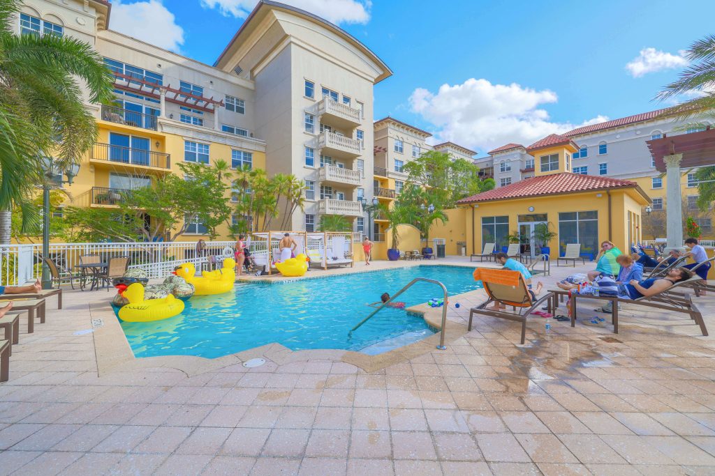 People relax by a sunny hotel pool. Some float on yellow inflatable toys, while others sit on lounge chairs. The surrounding buildings are multi-story with balconies. Palm trees and a clear blue sky complete the scene.