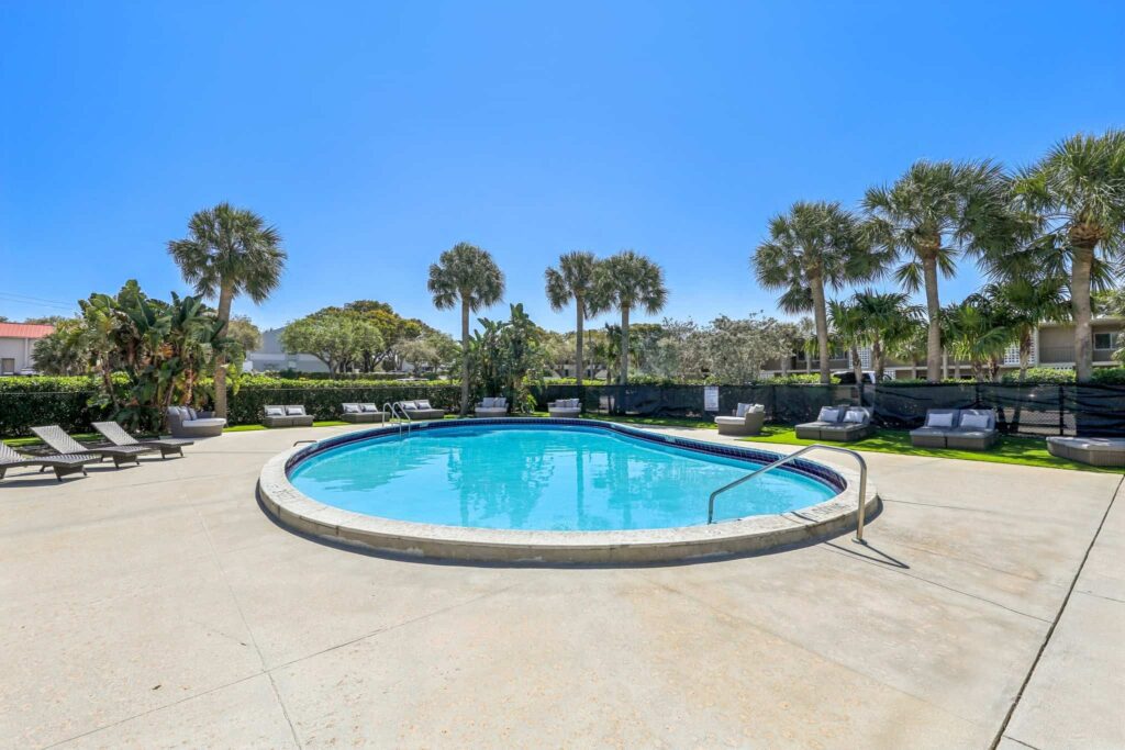 A serene swimming pool surrounded by lounge chairs on a sunny day. Palm trees and lush greenery are in the background under a clear blue sky.