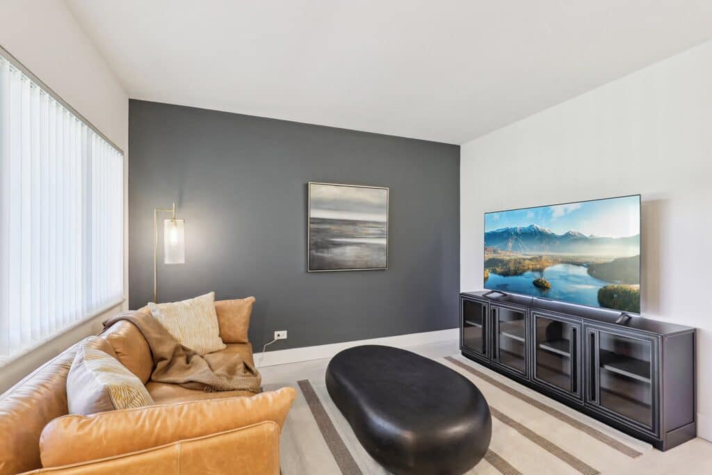 Modern living room with a brown leather sofa, pillows, and a throw blanket. Black oval coffee table on a striped rug. Large flat-screen TV on a black stand showing a scenic view. Dark accent wall with a framed abstract painting. Tall window with blinds.