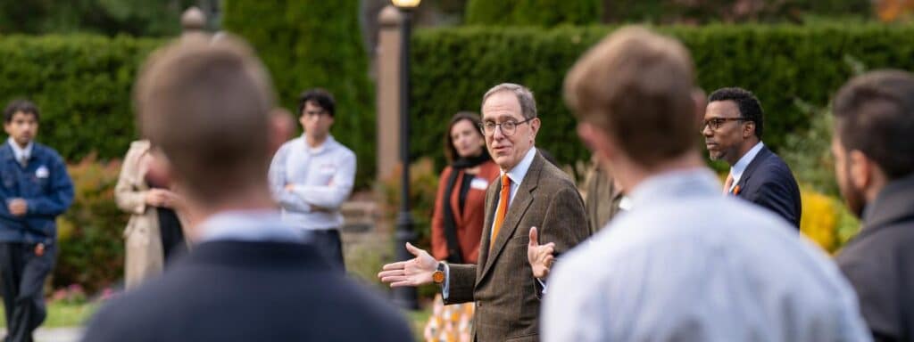 A group of people is gathered outdoors. A man in a suit and glasses is speaking, gesturing with his hands. Another man in a suit stands nearby. The background features greenery and a stone structure. Some people are blurred in the foreground.