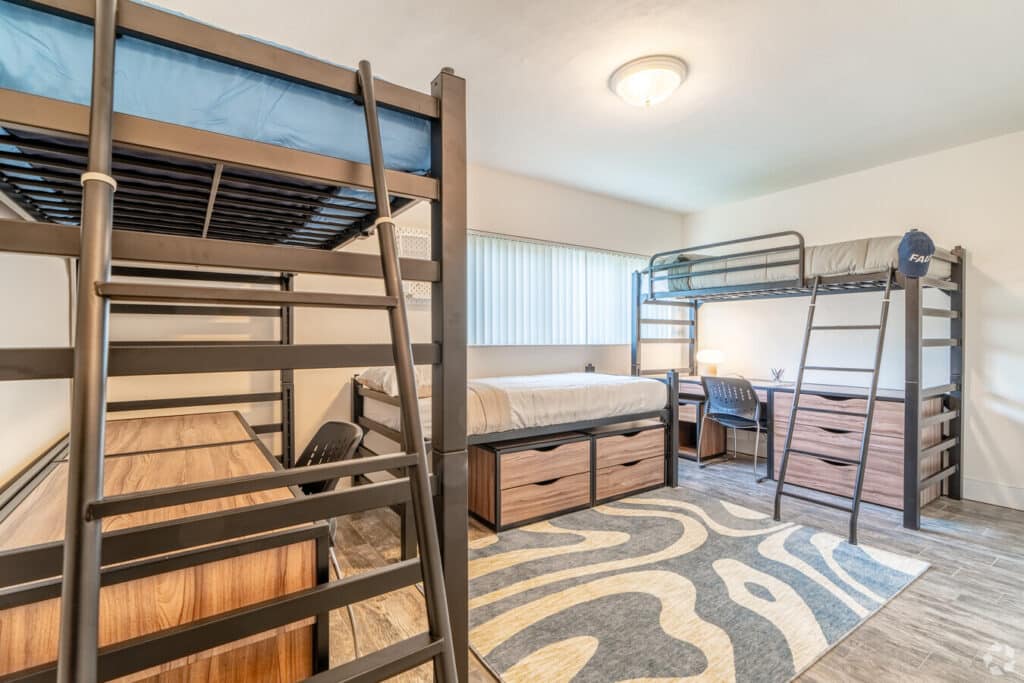 A dorm room with two lofted beds featuring wooden frames is perfect for FAU students. Each bed includes a study desk and drawers underneath. A patterned rug adorns the floor, and a window offers natural light. Ideal for off-campus living, each desk is complemented by a comfortable chair.