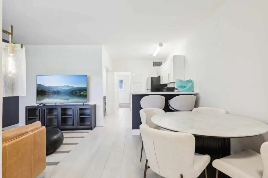 Modern living and dining area in Boca Raton Apartments for Rent, featuring a round marble table with four white chairs, a wall-mounted TV displaying a lake scene, a tan sofa, and a sleek kitchen with white cabinets in the background.
