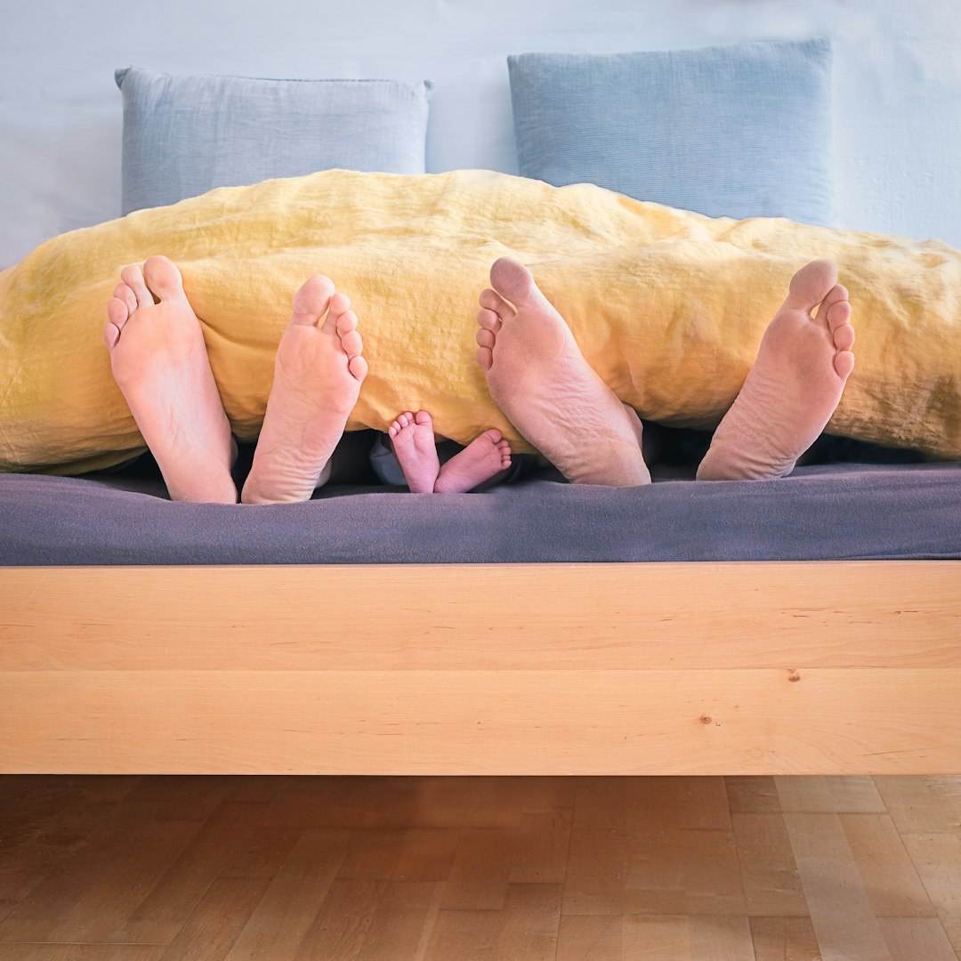 Three pairs of feet—two adults and one child—stick out from under a yellow blanket on a bed, suggesting a family lying together. Two pillows are visible in the background.